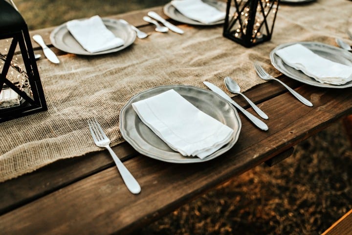 Picture of rustic wood table outdoors set with place settings and burlap table runner and rustic lanterns.